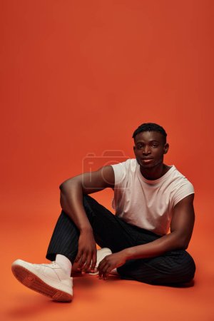 Afrikanisch-amerikanischer Mann in trendiger Freizeitkleidung sitzt und blickt in die Kamera auf rotem und orangefarbenem Hintergrund