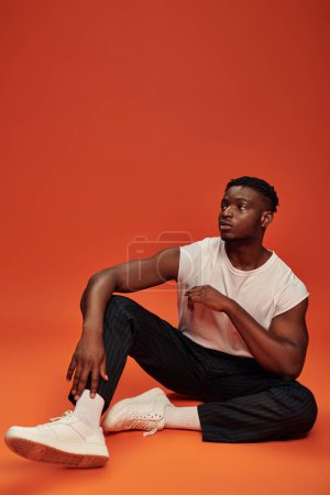 Photo for Full length of trendy african american guy sitting and looking away on red and orange backdrop - Royalty Free Image