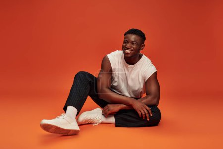 glücklich afrikanisch-amerikanischer Mann in stilvoller Streetwear sitzt auf rotem und orangefarbenem Hintergrund, volle Länge