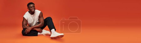 Téléchargez les photos : Pleine longueur de souriant homme afro-américain à la mode assis sur fond rouge et orange, bannière - en image libre de droit