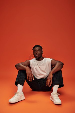Photo for Confident and sportive african american man sitting and looking at camera on red and orange backdrop - Royalty Free Image