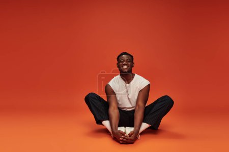Photo for Overjoyed african American man in white tank top and black pants sitting on red and orange backdrop - Royalty Free Image