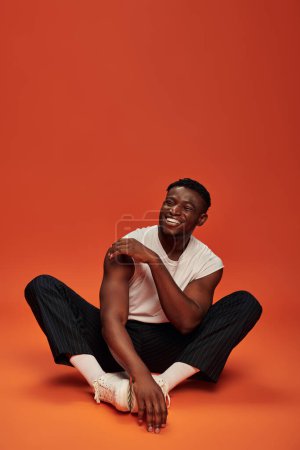 emocionado hombre afroamericano elegante sentado y riendo con los ojos cerrados, fondo rojo y naranja