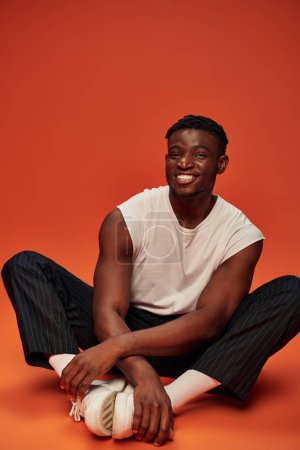 young african american male model in white tank top and pants sitting an smiling at camera on red