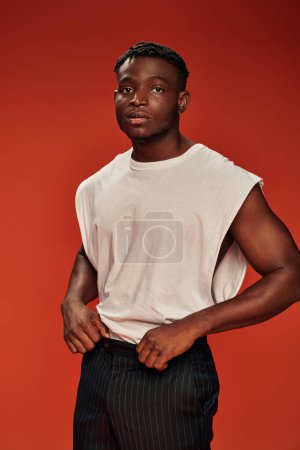pensive african american man in white tank top and black pants looking away on red backdrop