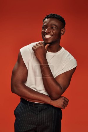 hombre afroamericano emocionado en ropa de calle elegante mirando hacia otro lado y riendo en rojo, vibraciones positivas
