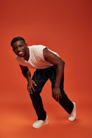 Foto de Chico afroamericano feliz en camiseta blanca y pantalones inclinados hacia adelante y mirando a la cámara en rojo - Imagen libre de derechos