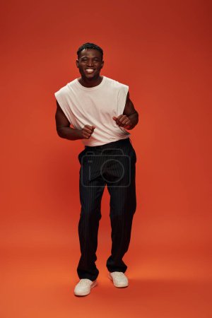 Foto de Hombre afroamericano lleno de alegría en ropa casual de moda posando sobre fondo rojo y naranja - Imagen libre de derechos