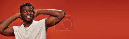 Photo for Smiley and hot african american guy standing with hands behind head and looking away on red, banner - Royalty Free Image