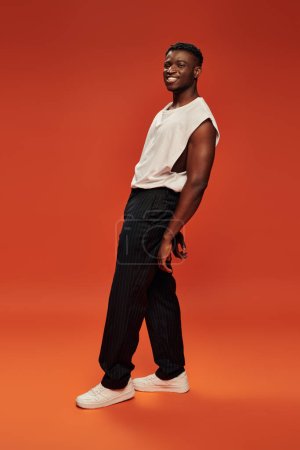 homme afro-américain insouciant en pantalon noir et débardeur blanc posant sur fond rouge et orange