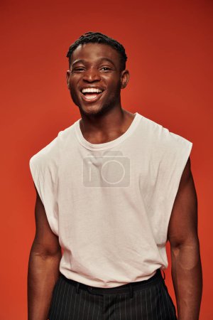 young excited african american male model in white tank top laughing and looking at camera on red