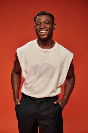 cheerful african american man with hands in pockets smiling at camera on red, hot and happy guy
