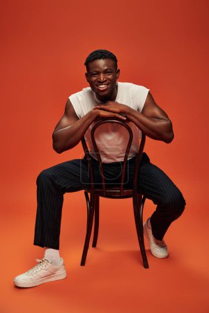 hombre afroamericano excitado en camiseta blanca y pantalones sentados en silla sobre fondo rojo y naranja