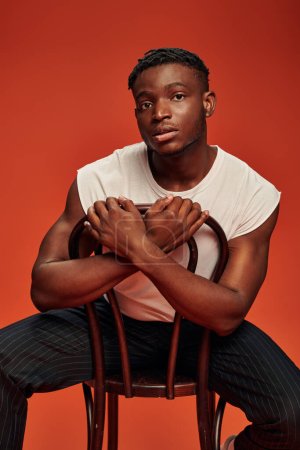 handsome african american man in white tank top sitting on chair and looking at camera on red