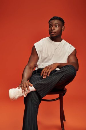 Foto de Chico afroamericano caliente en pantalones negros y camiseta blanca posando en la silla y mirando hacia otro lado en rojo - Imagen libre de derechos