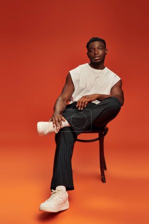 afrikanisch-amerikanisches Männermodel in stylischer Streetwear sitzt auf Stuhl vor rotem und orangefarbenem Hintergrund