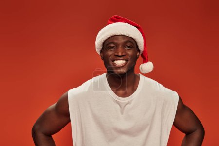 Foto de Alegre afroamericano hombre con sonrisa radiante posando en sombrero de santa y mirando a la cámara en rojo - Imagen libre de derechos