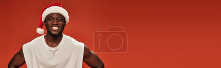 Photo for Joyful african american guy in santa hat and white tank top looking at camera on red, banner - Royalty Free Image