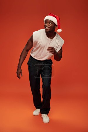 alegre afroamericano chico en santa hat y elegante casual ropa mirando hacia otro lado en rojo