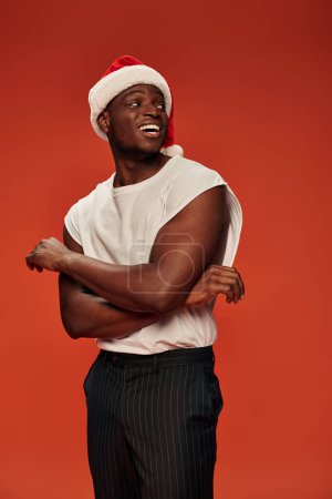 excited african american man in white tank top and christmas cap looking away on red backdrop