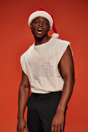 flirty african american man in santa hat and white tank looking at camera on red backdrop