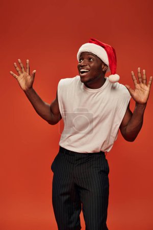 amazed and overjoyed african american man in santa hat showing wow gesture and looking away on red