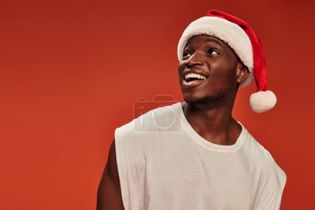 amazed and overjoyed african american man in santa cap smiling and looking away on red backdrop