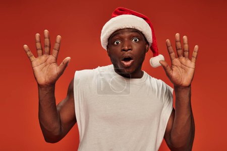 scared african american guy in christmas hat and with open mouth showing stop gesture on red