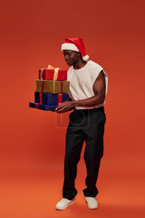 pleased african american man in festive santa hat holding colorful christmas presents on red