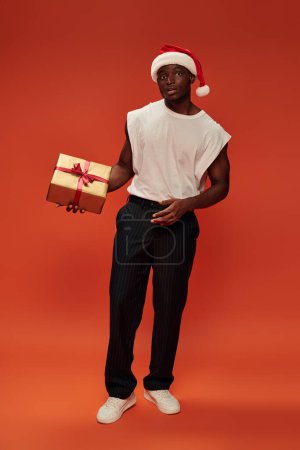 afrikanisch-amerikanischer Mann in trendiger Freizeitkleidung und Weihnachtsmütze mit goldener Geschenkschachtel auf rotem Grund, volle Länge