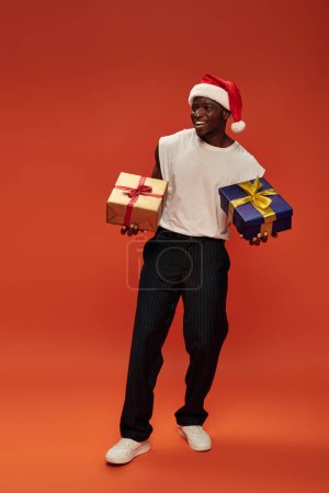 volle Länge der lustigen afrikanisch-amerikanischen Mann in lässiger Kleidung und Weihnachtsmann Mütze hält Geschenkboxen auf rot
