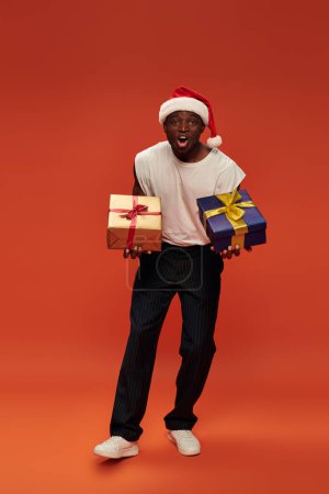 homme afro-américain excité en tenue décontractée et chapeau de Père Noël tenant cadeaux de Noël sur rouge