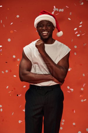 joyful african american man in christmas hat looking at camera on red backdrop with falling confetti