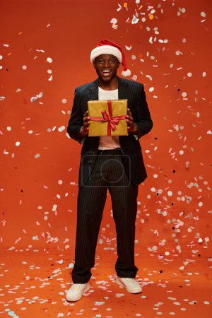 pleased dark skinned man in black suit and santa hat standing with gift box under confetti on red