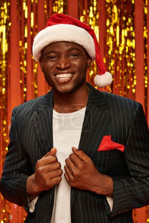 hombre afroamericano feliz en sombrero de santa y chaqueta negra sonriendo sobre fondo brillante con oropel