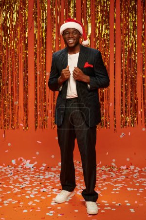 cheerful african american man in santa hat and black suit on orange backdrop with shiny decor