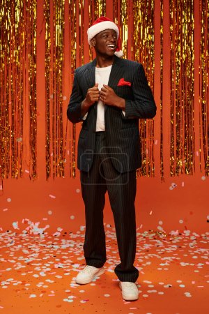 Photo for Excited african american man in santa hat and suit looking away near shiny tinsel on red backdrop - Royalty Free Image