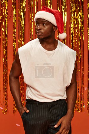 dark skinned guy in santa hat holding hands in pockets and looking away near golden tinsel on red