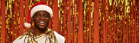 cheerful african american man in santa cap smiling at camera near shiny tinsel on red, banner