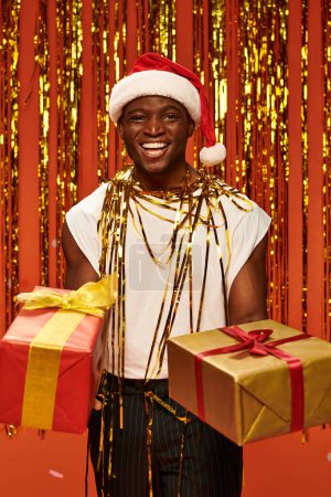 Foto de Hombre afroamericano lleno de alegría en santa gorra con regalos de Navidad cerca de oropel de oro en rojo - Imagen libre de derechos