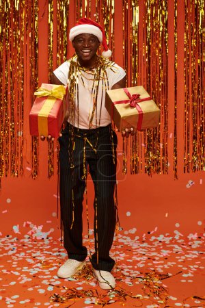 Afrikanisch-amerikanischer Mann in Weihnachtsmütze mit Geschenken und Blick in die Kamera in der Nähe von Weihnachtsdekor auf rot