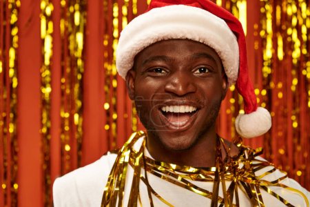 portrait of excited african american guy in santa hat laughing near golden tinsel on red backdrop