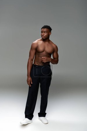 longitud completa de hombre afroamericano musculoso y sin camisa en pantalones negros mirando hacia otro lado en gris