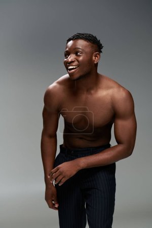 Foto de Emocionado y sin camisa chico afroamericano con cuerpo fuerte riendo y mirando hacia otro lado en gris - Imagen libre de derechos