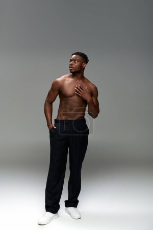 longitud completa de hombre afroamericano sin camisa y musculoso con la mano en bolsillos mirando hacia otro lado en gris