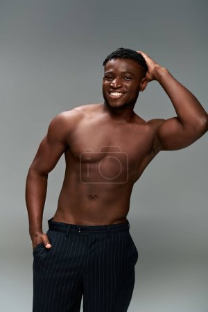 athletischer afrikanisch-amerikanischer Mann mit hemdlosem Körper und Hand in Tasche, der in grau in die Kamera lächelt