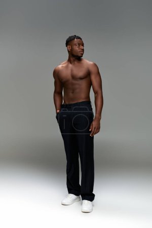 sportlicher afrikanisch-amerikanischer Mann mit kräftigem Körper und einer Hand in der Hosentasche, der wegschaut auf grau, volle Länge