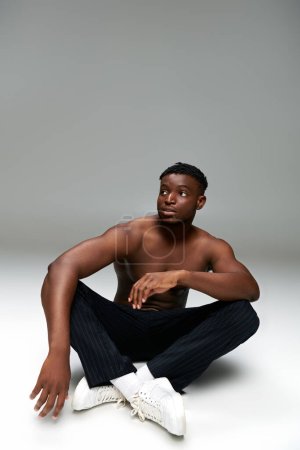 Foto de Sin camisa atlético africano americano hombre en pantalones negros sentado y mirando hacia otro lado en gris, longitud completa - Imagen libre de derechos