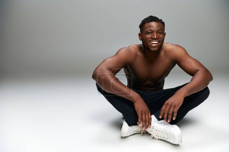 fröhlicher afrikanisch-amerikanischer Typ mit starkem, hemdlosen Körper, der auf grauen Ganzkörperanzügen sitzt und lächelt