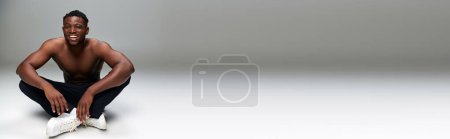 Photo for Joyful african american guy with strong shirtless torso sitting and smiling on grey backdrop, banner - Royalty Free Image